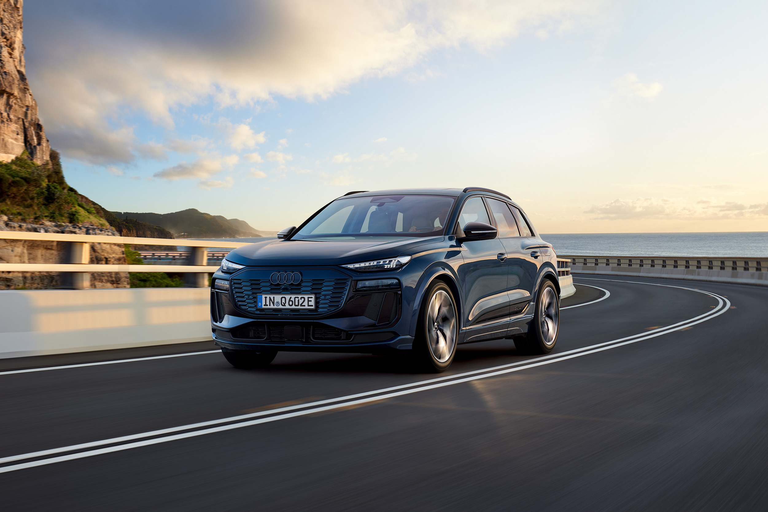 Ein fahrender Audi Q6 SUV e-tron in Front-Seitenansicht mit idyllischer Meereskulisse im Hintergrund